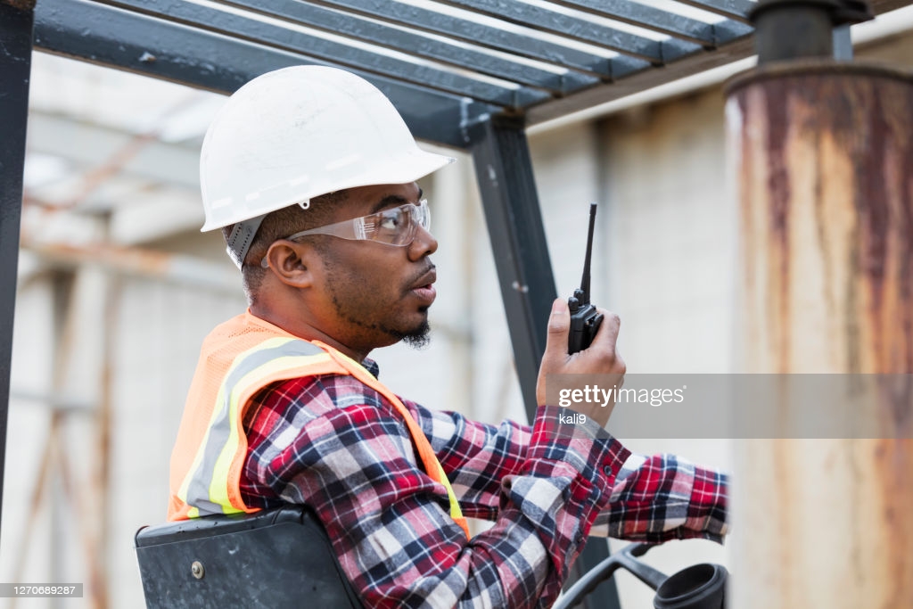 A mid adult African-American man in his 30s operating a forklift outside a warehouse. He is wearing a hard hat, safety glasses, reflective vest and plaid shirt, sitting in the driver's seat, talking on a walkie-talkie.